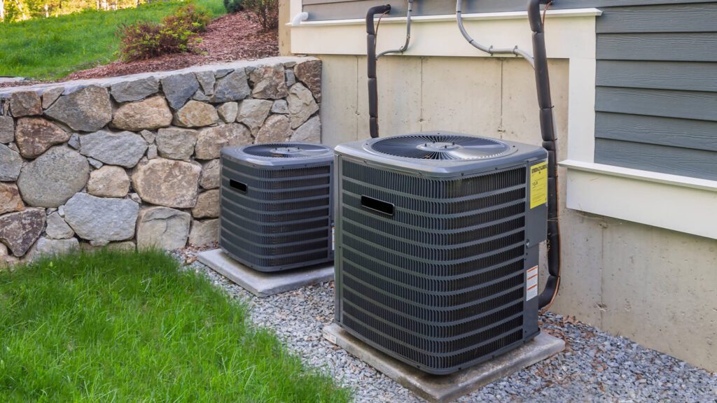 Two outdoor air conditioning units on gravel pads beside a house